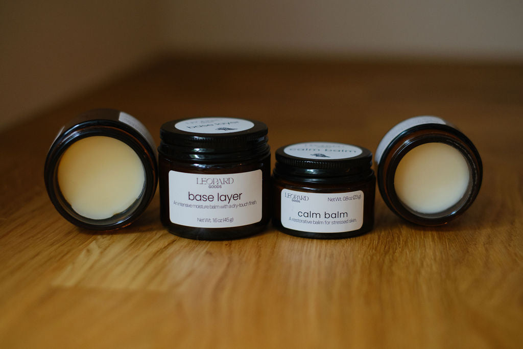 Jars of Base Layer and Calm Balm, both open and closed, on a wooden surface