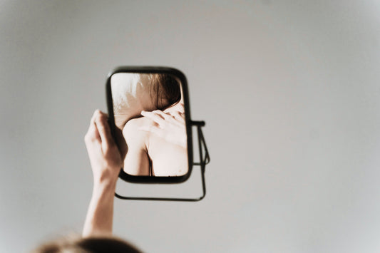 Woman holding up mirror looking at shoulder with gray wall behind