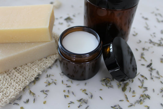 Skin balm in an amber jar on a white background with soap, a loofah, and plant material.