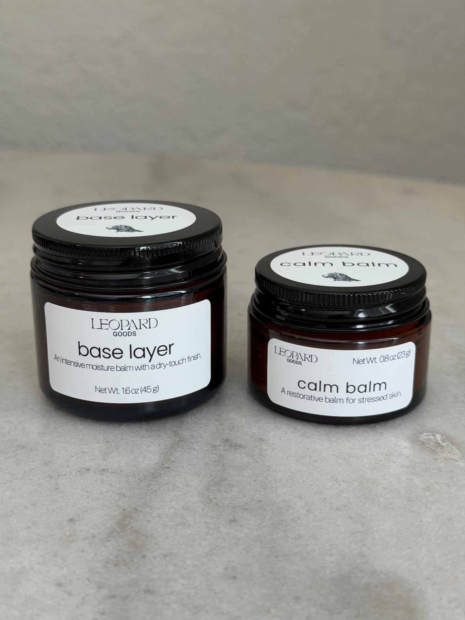 Two jars of Leopard Goods skincare, Base Layer, and Calm Balm, on a gray marble surface